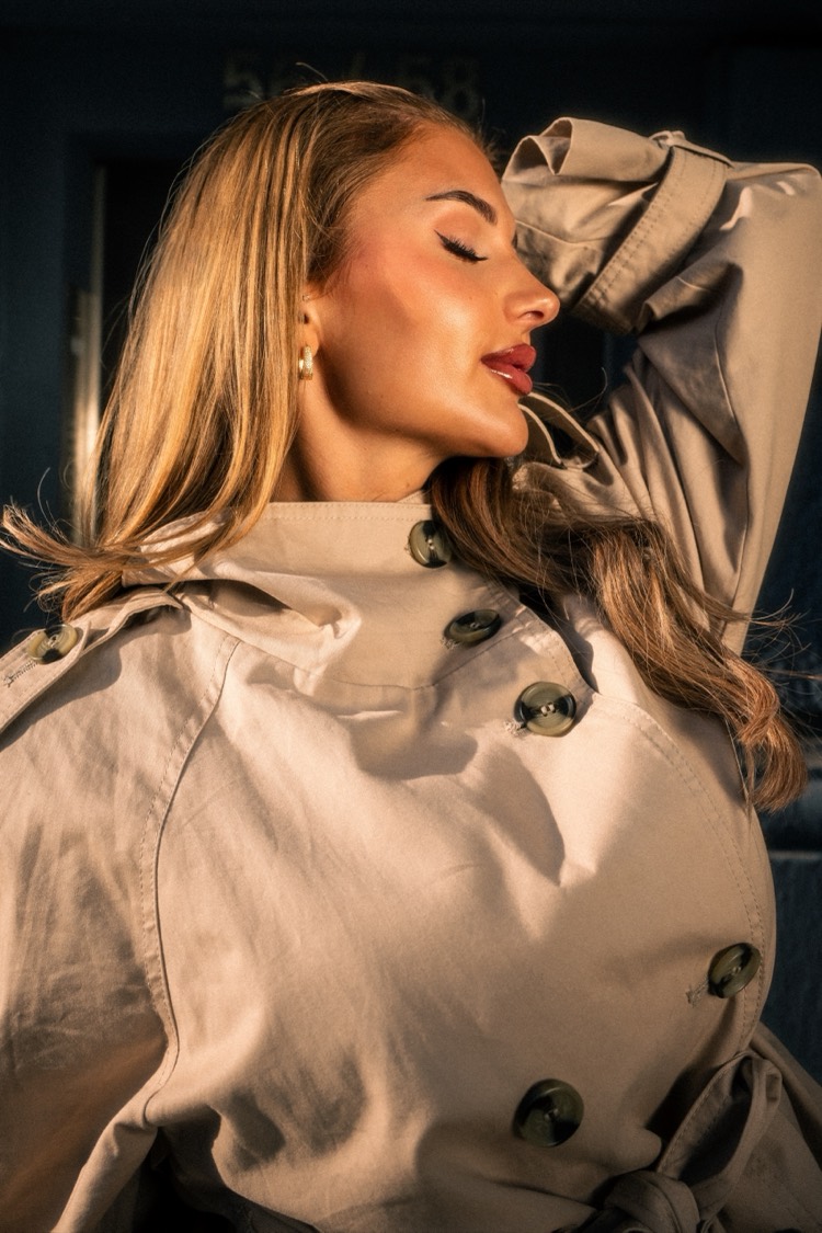 Nahaufnahme einer jungen Frau mit langem blondem Haar im beigen Trenchcoat, Augen geschlossen, Haar im Wind, tiefe Abendsonne.