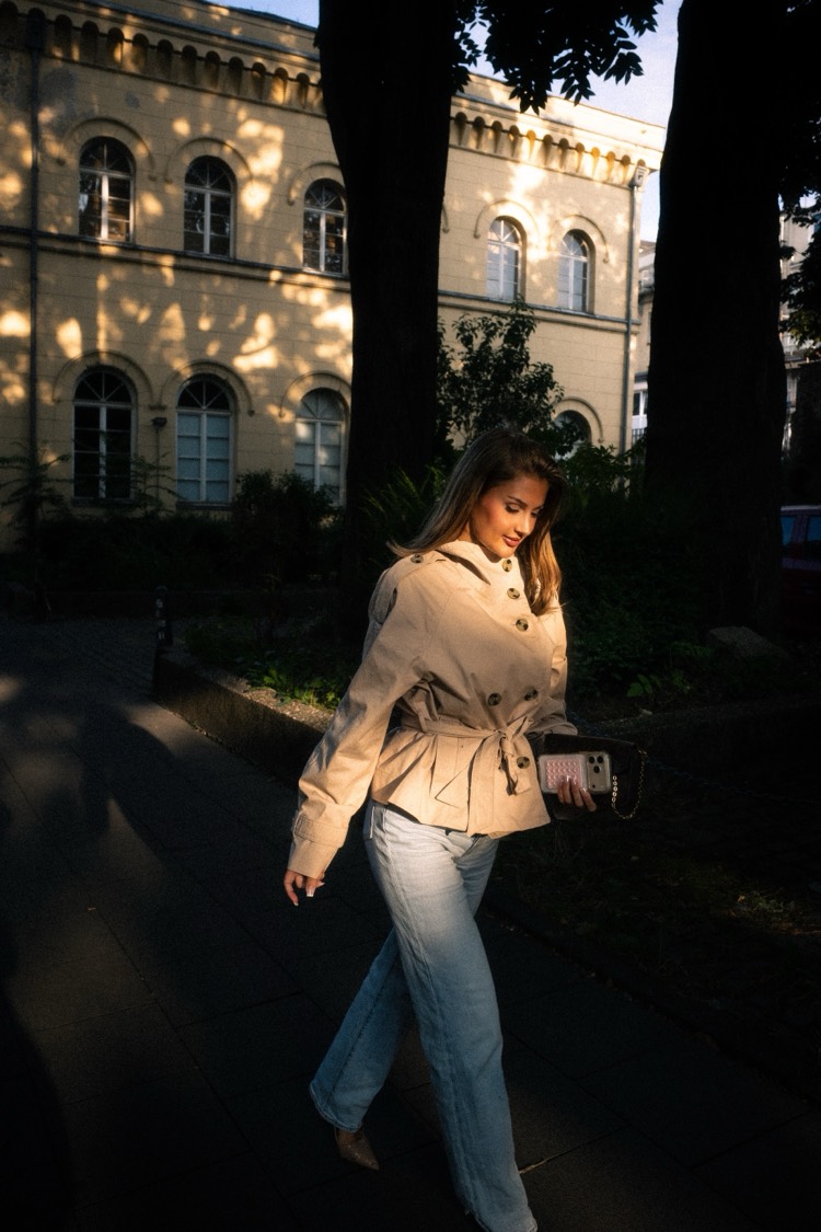Junge Frau im beigen Trenchcoat geht mit Tasche durch Streifen aus Abendsonne, dahinter Altbau und Bäume.