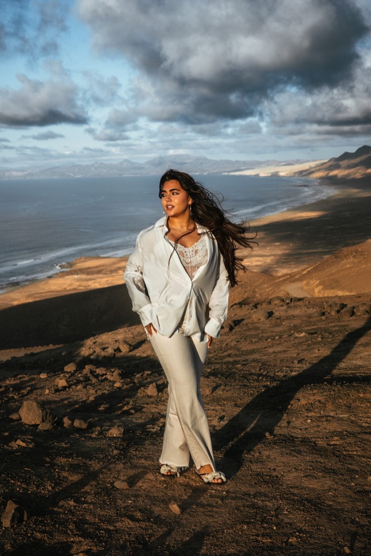 Junge Frau in weißer Bluse und heller Hose steht auf einem Vulkanhang über dem Meer, Haar im Wind, Abendlicht auf Dünen und Bergen.
