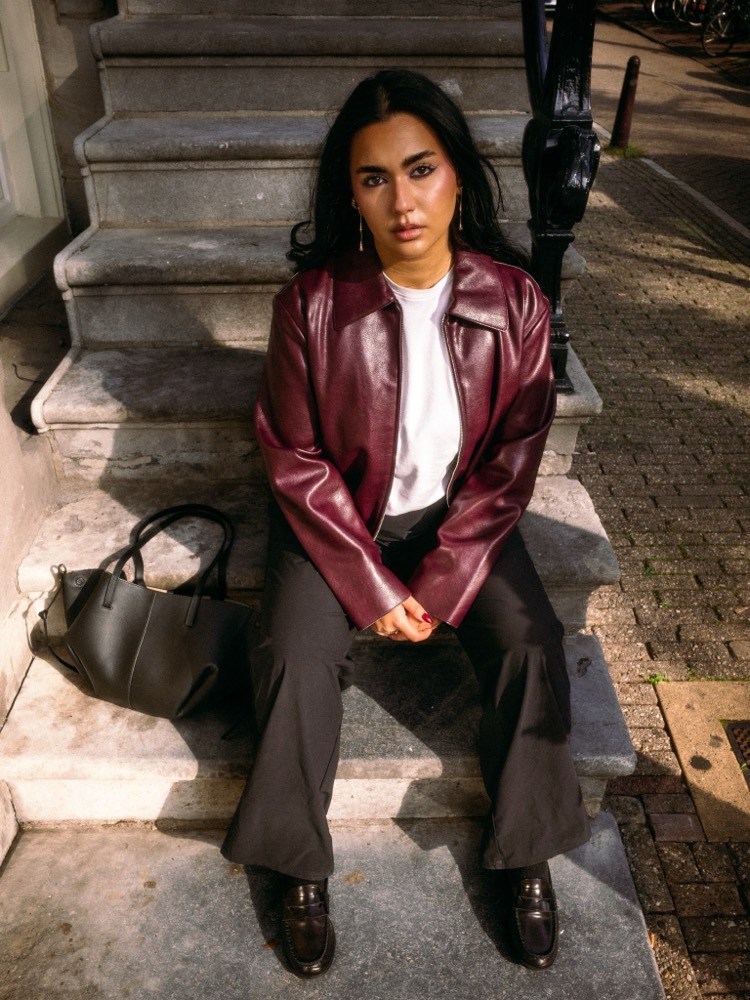 Junge Frau in weinroter Lederjacke und weißem Shirt sitzt frontal auf einer Steintreppe, eine Tasche neben sich, Blick in die Kamera.