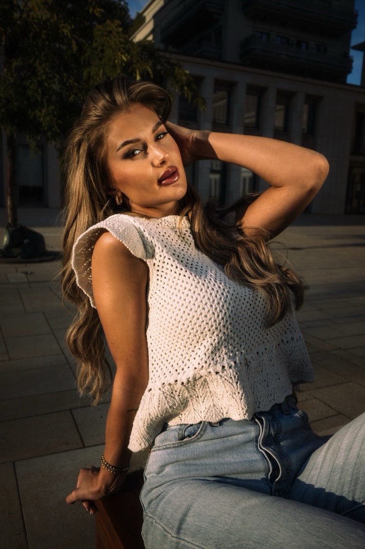 Junge Frau mit langem, gewelltem Haar in weißem Häkeltop und heller Jeans sitzt in der Abendsonne auf einer Bank, eine Hand im Haar, dahinter ein Baum und eine Bürofassade.
