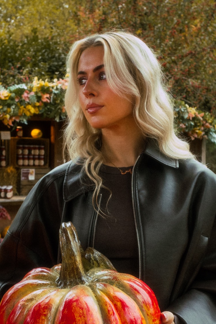 Nahaufnahme einer jungen Frau mit hellblondem Haar in schwarzer Lederjacke, sie blickt zur Seite, vorn ein großer bemalter Kürbis, dahinter Blumen und Regale.