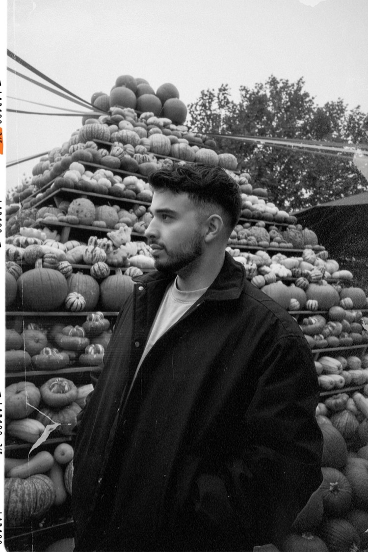 Porträt in Schwarzweiß im Profil: kurzer Bart, dunkle Jacke über hellem Shirt, dahinter eine Pyramide aus Kürbissen und Bäume.